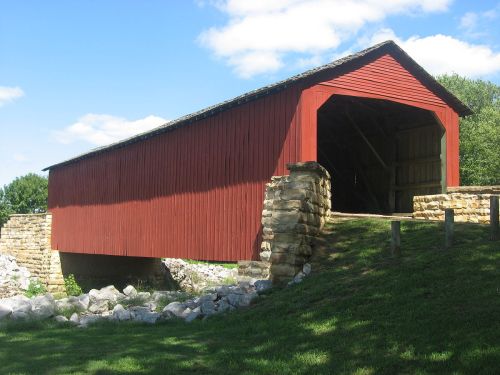 A red covered bridge with stone supports, set against a blue sky and green grass.
