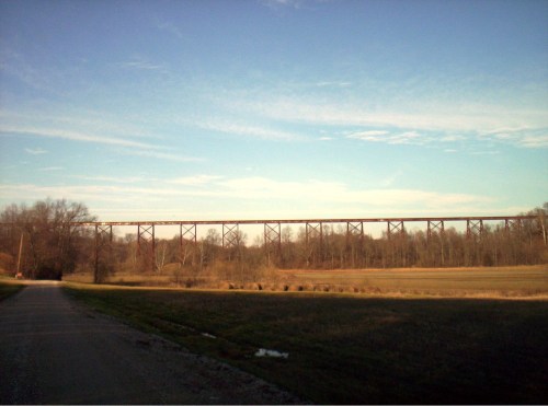 A tall, rusty bridge spans a rural landscape under a clear blue sky, with a dirt road leading towards it.