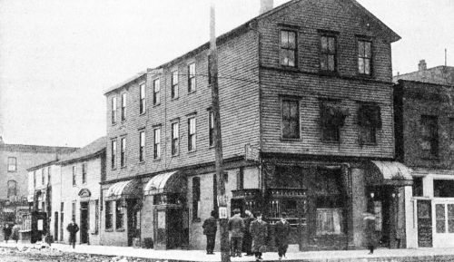 Historic black-and-white photo of a three-story wooden building with storefronts, people gathered outside on the street.