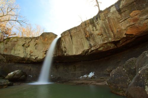A serene waterfall cascading over a rocky cliff into a calm pool, surrounded by trees and a clear blue sky.