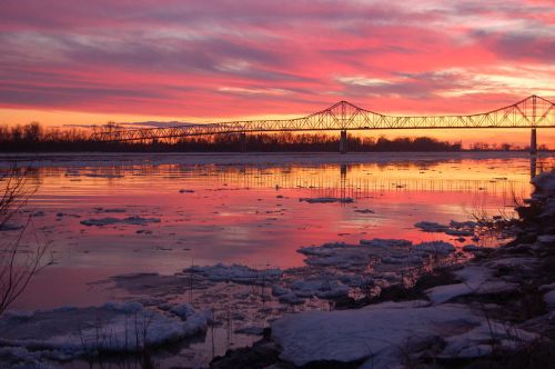 A vibrant sunset reflects on a river with ice chunks, framed by a bridge and silhouetted trees in the background.
