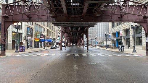 Empty city street beneath an elevated train track, with buildings lining the sides and no pedestrians or vehicles in sight.