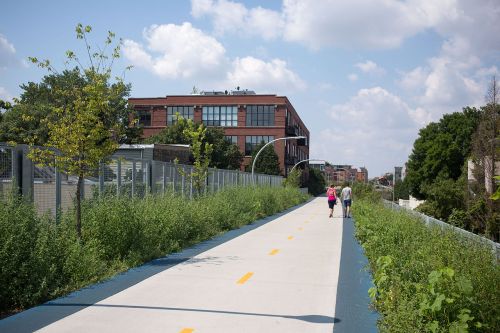 A paved path lined with greenery, where two people walk side by side, with a building visible in the background.