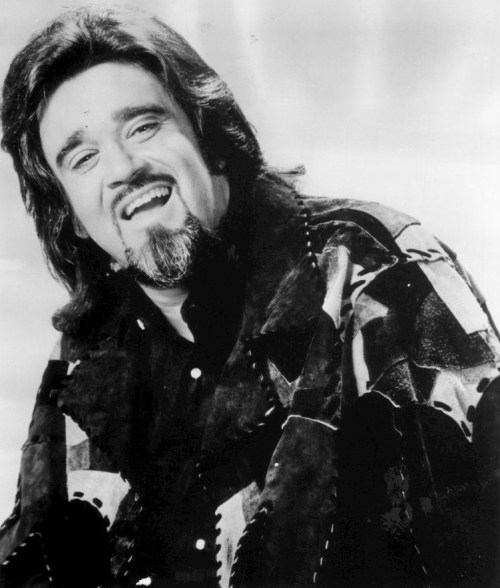 A smiling man with long hair and a patterned jacket, captured in a black and white portrait.