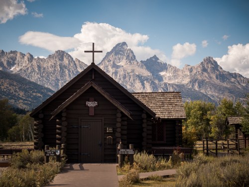 A rustic log chapel with a cross, set against a backdrop of majestic mountains and a clear blue sky.