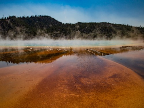 A vibrant geothermal pool with orange and blue hues, surrounded by mist and forested hills in Yellowstone National Park.