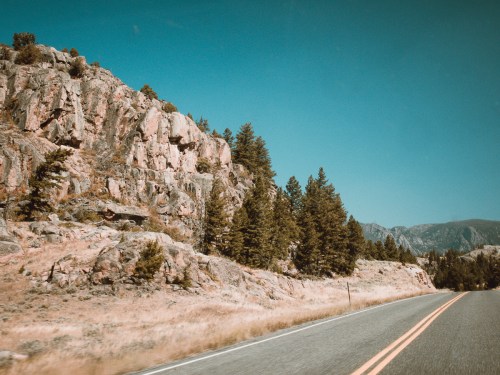 A scenic road lined with rocky cliffs and pine trees under a clear blue sky.