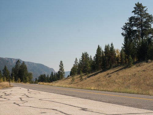 A cracked road stretches alongside a grassy hillside and trees, with mountains in the background under a clear blue sky.