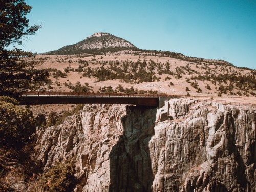 A bridge spans a rocky cliff with a mountain in the background under a clear blue sky.