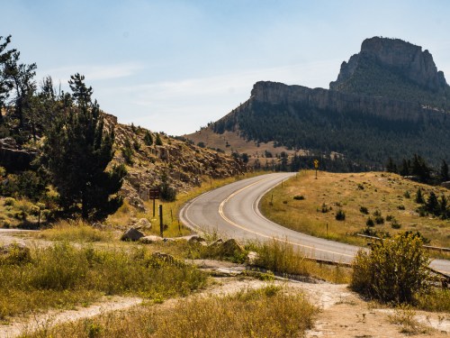 A winding road leads through a grassy landscape with a rocky mountain in the background under a clear blue sky.