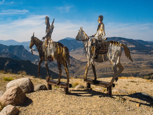Two metallic sculptures of riders on horseback stand against a scenic mountain backdrop under a clear blue sky.