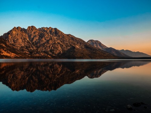 A serene lake reflects majestic mountains under a clear blue sky at sunset.
