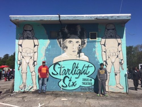 Mural on a building featuring a woman with stormtroopers, with two people standing in front at a drive-in theater.