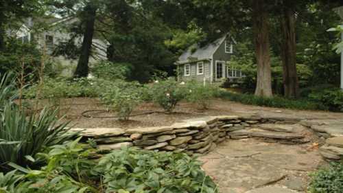 A serene garden with a stone pathway leading to a charming house surrounded by trees and greenery.