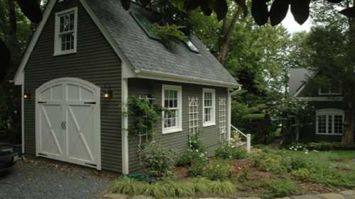 A charming gray house with white trim, surrounded by greenery and flowers, featuring a gabled roof and double doors.