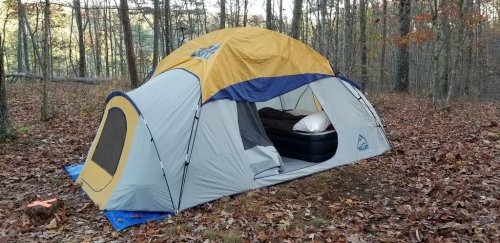 A large tent set up in a wooded area, surrounded by fallen leaves and trees, with an air mattress visible inside.
