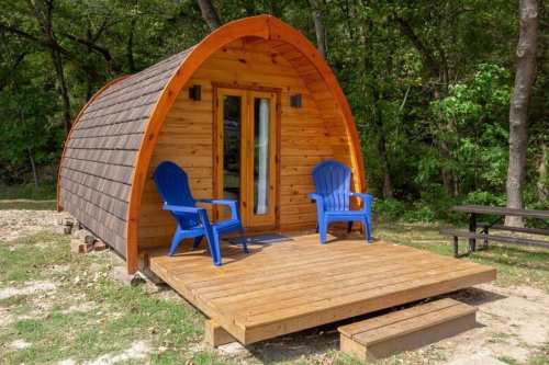 A cozy wooden cabin with a curved roof, featuring two blue chairs on a wooden deck surrounded by greenery.