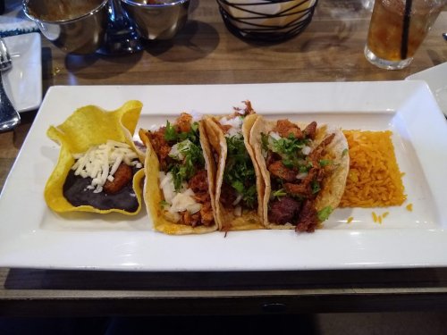 A plate with three tacos filled with meat and toppings, served with rice and a side of black beans in a tortilla shell.