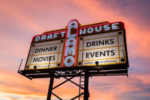 Sign for Alamo Drafthouse featuring "Dinner," "Movies," "Drinks," and "Events" against a colorful sunset sky.