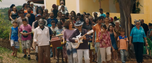 A diverse group of people walking together, some carrying instruments, in a vibrant outdoor setting.
