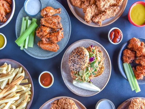 A spread of fried chicken, wings, fries, a sandwich, celery sticks, and various dipping sauces on a dark table.