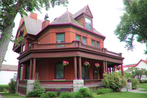 Victorian-style house with a large porch, decorative trim, and colorful flower baskets, surrounded by greenery.