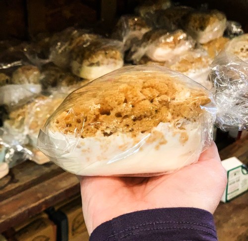 A hand holding a wrapped dessert with a crumbly topping, surrounded by more similar treats in the background.