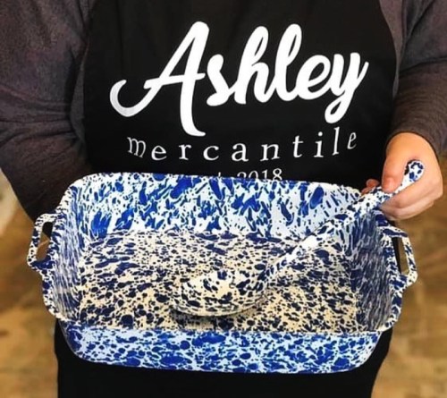 A person in an apron holds a blue and white speckled serving dish and spoon. The apron features the name "Ashley."