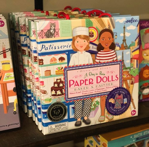 A stack of colorful paper doll books titled "A Day in Paris" featuring characters in baker and painter outfits.