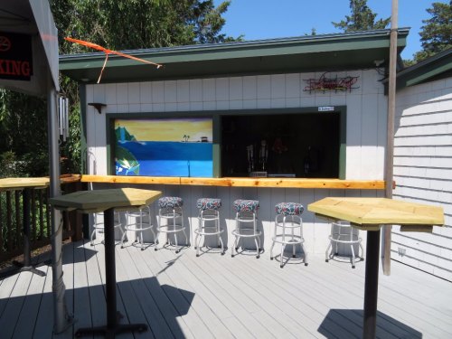 Outdoor bar area with wooden tables, colorful stools, and a mural of a beach scene on the wall.
