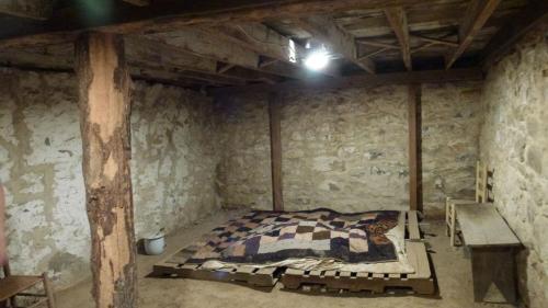 A dimly lit, rustic room with stone walls, a wooden beam, a quilt on a pallet, and simple wooden furniture.