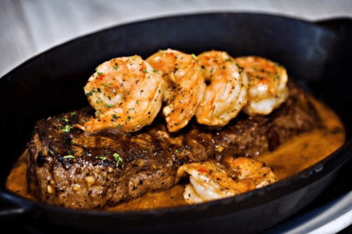 A juicy steak topped with grilled shrimp, garnished with herbs, served in a black skillet.