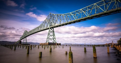 A large green bridge spans over calm waters, with wooden pilings visible beneath and a cloudy blue sky above.