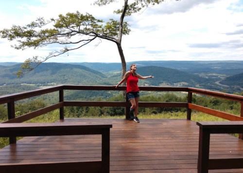 A woman in a red tank top stands on a wooden deck, gesturing towards a scenic mountain view under a cloudy sky.