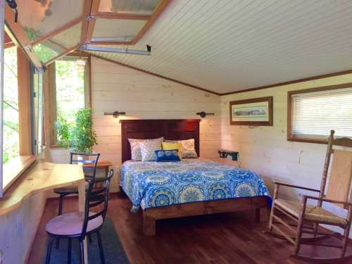 Cozy bedroom with a wooden bed, blue patterned bedding, a rocking chair, and large windows letting in natural light.