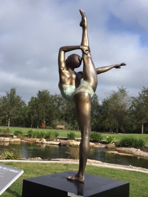 A bronze sculpture of a dancer in a graceful pose, set in a landscaped park with trees and a pond in the background.