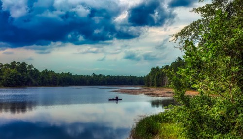 A serene lake scene with a lone kayaker, surrounded by lush greenery and dramatic clouds overhead.
