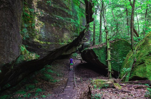 A hiker walks along a narrow path between large moss-covered boulders in a lush green forest.