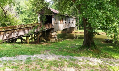 A rustic covered bridge spans a small pond, surrounded by lush greenery and trees.