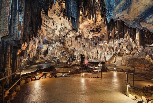 A vast cave interior with stalactites and stalagmites, illuminated pathways, and rocky formations.