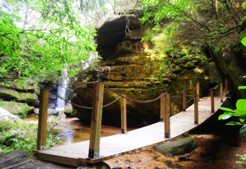 A wooden bridge with a chain railing crosses a serene stream, surrounded by lush greenery and rocky cliffs.