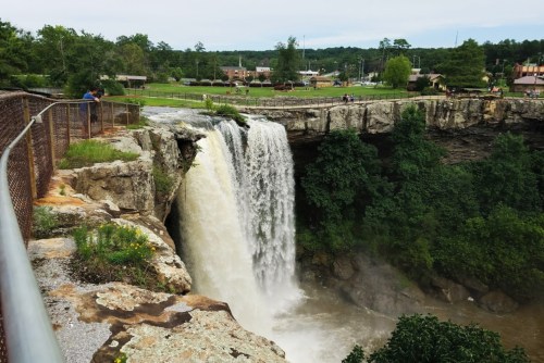 A waterfall cascades over a rocky ledge, surrounded by greenery and a viewing area with visitors nearby.