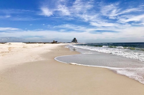 A serene beach scene with gentle waves, soft sand, and a distant skyline under a blue sky with wispy clouds.