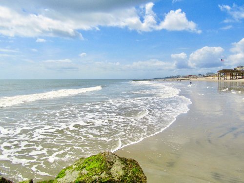 A serene beach scene with gentle waves, a rocky foreground, and a distant pier under a partly cloudy sky.