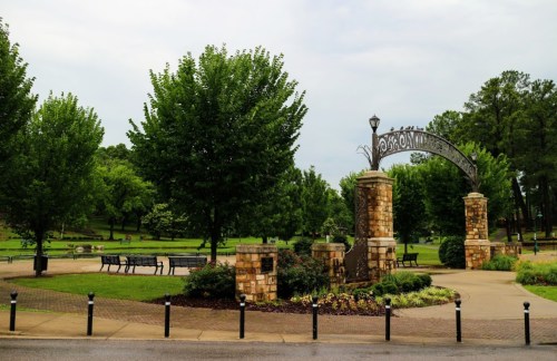 Entrance to a park with stone pillars, greenery, benches, and a cloudy sky in the background.