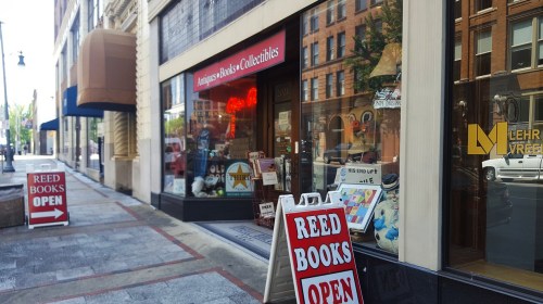 A street view of a bookstore with a sign reading "Reed Books Open" and collectibles displayed in the window.