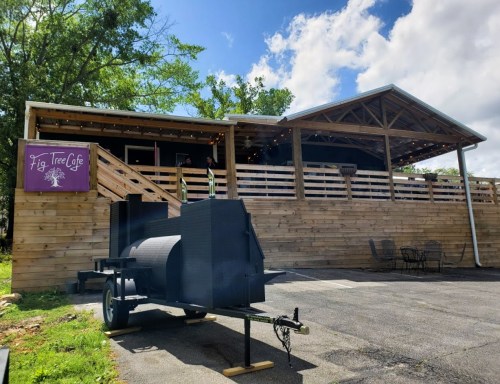 A wooden café with a sign reading "Fig Tree Café," featuring a smoker outside and a deck with seating.
