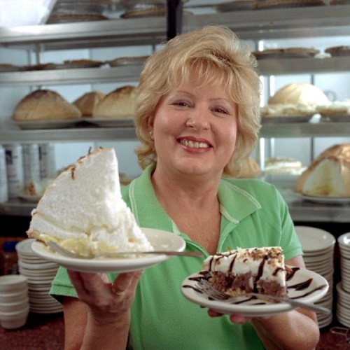 A smiling woman holds a slice of coconut cream pie and a plate of chocolate dessert in a bakery setting.