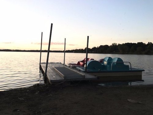 A calm lake at sunset with a wooden dock and paddle boats moored nearby.