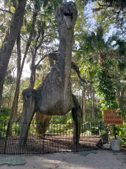 A large dinosaur statue surrounded by trees, with a sign that reads "DO NOT FEED THE DINOSAURS."